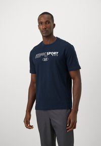 T-shirt de algodão azul-marinho com mangas curtas, apresentando o texto branco "ICONIC SPORT READY SET MOVE" na parte frontal, com decote redondo e corte standard.