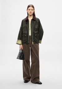 Tume roheline vahaine jope pruunide kraega, kuldsete detailide ja kahe eesmise taskuga. Modell kannab pruunid laia säärega pükse ning hoiab käes väikest musta kotti.