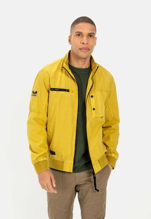 Yellow utility jacket with a zip and snap closure, featuring a chest pocket, elastic cuffs, and side pockets; worn over a green t-shirt.