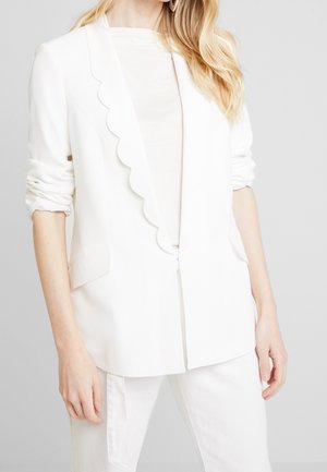 Mujer con blazer blanco a medida con bordes festoneados en la solapa, top blanco y pantalones blancos sobre un fondo gris claro y liso.
