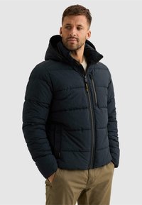 Homme portant une veste matelassée bleu marine foncé avec une capuche et les mains dans les poches, regardant sur le côté, debout devant un fond uni.