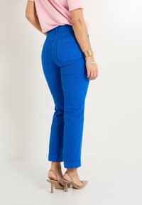 Jeans taperos de un azul brillante con cintura alta, que presentan dos bolsillos traseros y puños doblados. Combinados con sandalias de tacón de color beige claro.