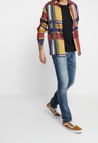 Rutig button-up skjorta i gult, rött, lila och blått, i kombination med en svart t-shirt, blå jeans och bruna mockaskor.