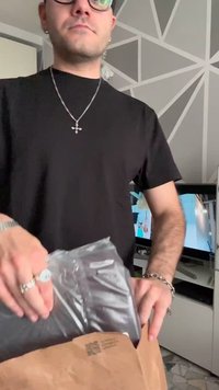 Man in zwart T-shirt en bril haalt een zwarte plastic verpakking uit een bruine envelop in een kamer met geometrische wandkunst.