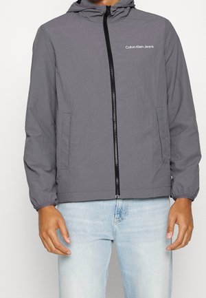 Mand iført en grå hættejakke med lynlås foran og sidelommer, lyseblå jeans, Calvin Klein Jeans-logo på brystet.