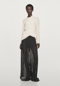 Cream blouse with a high neck and fitted waist, paired with sheer black pleated trousers, showcasing fluid texture and movement.