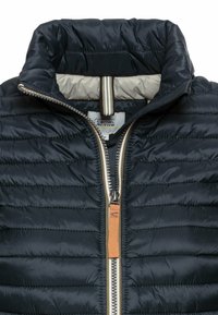 Schwarze gesteppte Jacke mit beigem Innenfutter, hohem Kragen und beigem Reißverschlussbesatz mit orangefarbenem Zugband.