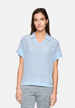 Frau mit langen braunen Haaren, die eine hellblaue Bluse mit kurzen Ärmeln und gemusterte Hosen trägt, vor einem schlichten weißen Hintergrund steht.