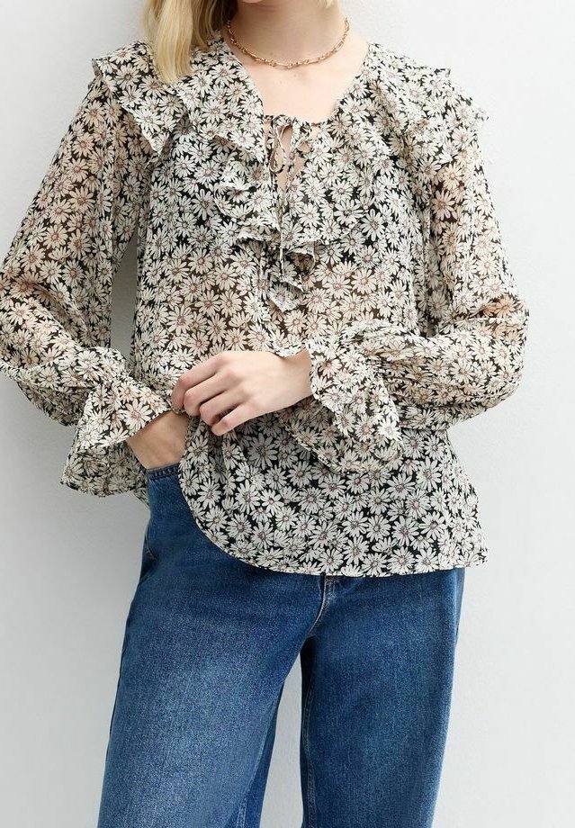 Blouse fleurie et transparente avec volants au niveau du col et des poignets, comprenant un détail à lacets. Fond noir avec motif floral blanc. Coupe élégante et texturée.