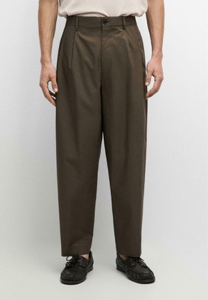 Mężczyzna w brązowych, plisowanych spodniach z wysokim stanem, czarnych sznurowanych butach i wpuszczonej w spodnie beżowej koszuli, stojący na jednolitym tle.