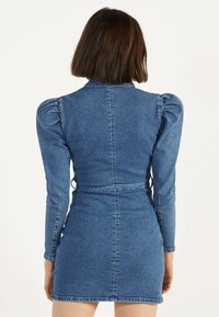Vestido de denim con una silueta ajustada, mangas largas abullonadas y cintura ceñida con cinturón. El material es de un azul medio con una textura de denim.