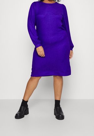 Fioletowa sukienka z długim rękawem o miękkim, teksturowanym wykończeniu. Projekt ma okrągły dekolt i luźny krój, zestawiona z masywnymi czarnymi butami.