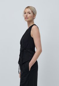 Svart ermeles topp med knytting i livet, med V-hals og jevn tekstur; designet for en tettsittende silhuett.