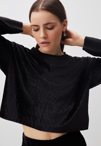 Schwarzes, kurzes Oberteil aus strukturiertem Stoff mit langen Ärmeln und rundem Halsausschnitt. Das Design zeigt subtile Wellen für zusätzliche Details.