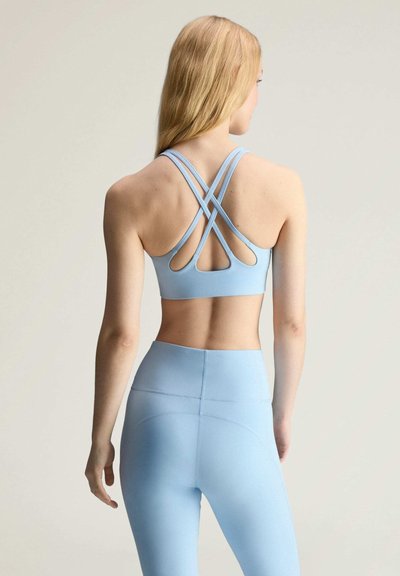 Woman with long blonde hair wearing light blue crisscross strap sports bra and matching high-waisted leggings, shown from behind.