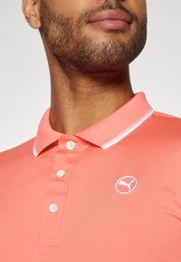 Polo shirt color corallo realizzato in tessuto testurizzato, con colletto a righe bianche, tre bottoni e logo Puma bianco sul petto.