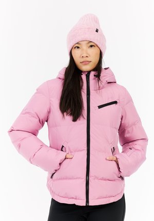 Puffige rosa Jacke mit Kapuze, Reißverschluss vorne und schwarzen Akzenten. Strukturierte gestrickte rosa Mütze. Modell posiert mit den Händen in den Taschen, neutraler Hintergrund.