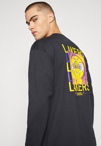 Juodas ilgomis rankovėmis marškinėliai su Los Andželo Lakers grafika purpurine ir geltona spalvomis nugaros pusėje, pagaminti iš medvilnės.
