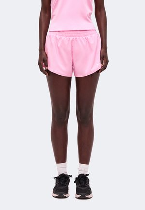 Person iført lyserøde træningsshorts og tanktop, hvide sokker og sorte løbesko, stående mod en ensfarvet baggrund.