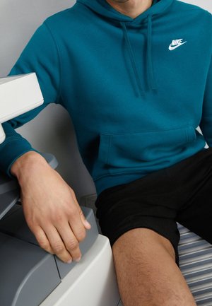 Osoba ubrana w turkusową bluzę z kapturem Nike i czarne spodenki, siedząca z jedną ręką opartą na szarej maszynie, ukazująca część tułowia i nóg.