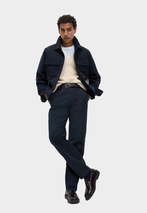 Man draagt een donkerblauw jasje, een crèmekleurig sweatervest over een wit overhemd, donkere broek en donkerleren loafers, staande met gekruiste benen.