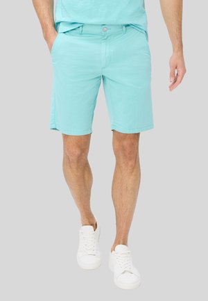 Mand iført turkis shorts og hvide sneakers, står med den ene hånd i lommen mod en ensfarvet baggrund.