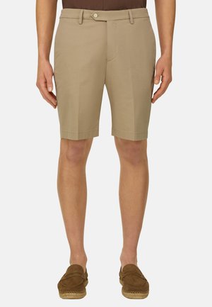 Beige maßgeschneiderte Shorts, getragen mit braunen Slipper-Loafern, gezeigt von der Taille bis zu den Füßen einer stehenden Person vor weißem Hintergrund.