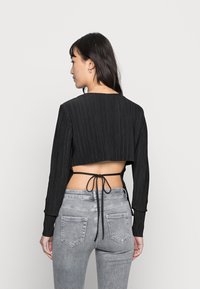 Top cropped preto com textura em nervuras verticais, mangas longas e detalhe de laço nas costas. Combinado com jeans justos cinza claro com bolsos traseiros.