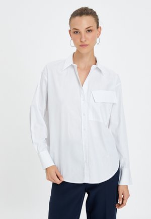 Camicia bianca con bottoni, caratterizzata da maniche lunghe, una tasca sul petto e un orlo curvo. Realizzata in tessuto leggero e morbido.