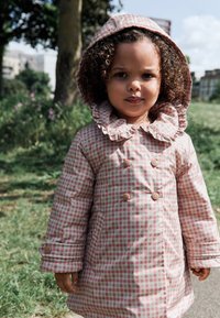 Imperméable à motif vichy rose et blanc avec une capuche à volants, des boutons croisés et un tissu texturé. Dispose de manches longues et de poches.
