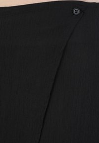 Tissu noir côtelé avec une texture douce, doté d'un bouton sur le côté et d'un design superposé. Le matériau est léger et ajusté.