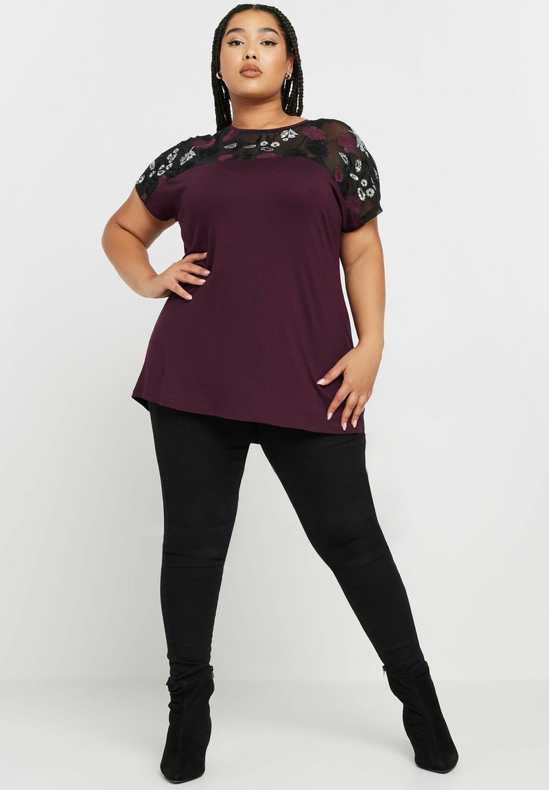 Burgundin t-paita, jossa on läpikuultavat mustat kukkakirjailtu lyhyet hihat, yhdistetty mustiin legginsseihin ja polveen asti ylettäviin mustiin saappaisiin. Silkkinen pinta.