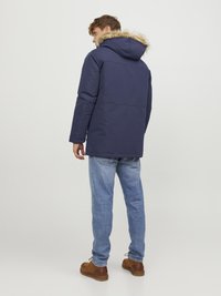 Parka blu navy con cappuccio foderato in pelliccia sintetica, maniche lunghe, chiusura con zip e tasche. Abbinato a jeans azzurri e scarpe marroni.