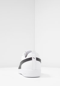 Biała sneaker z czarnym paskiem po bokach i logo Puma na zadniej części, sfotografowana z tyłu na białej powierzchni i tle.