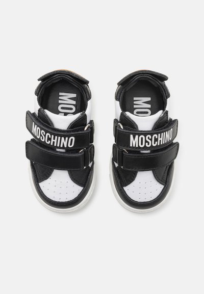 Sneakers bianco e nero con una punta forata, doppie strap in Velcro con il testo "MOSCHINO" e una suola bianca testurizzata.