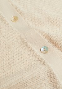 Gros plan sur un tissu tricoté beige avec un motif texturé et deux boutons ronds brillants sur une patte de boutonnage lisse.