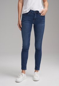 Jeans skinny blu scuro a vita media, con cinque tasche e una leggera scoloritura, abbinati a sneakers bianche con lacci.