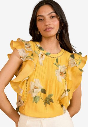 Geel bloemenblouse met gefronste mouwen, plooien en ingewikkelde kanten afwerking langs de halslijn, benadrukt door een botanisch patroon.
