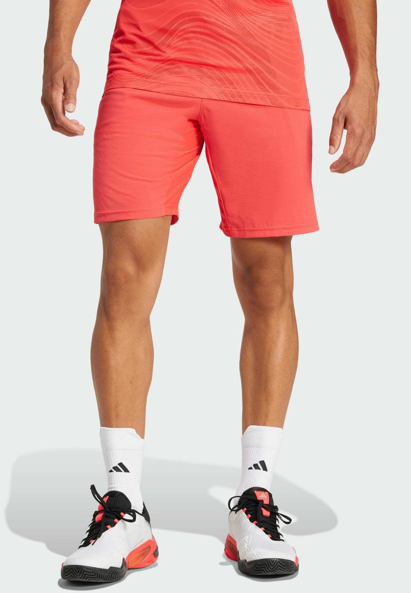 Shorts de sport pour hommes en corail, en mélange de polyester, texture lisse, longueur genou, associés à des chaussettes blanches et des chaussures noir-rouge, avec un logo.