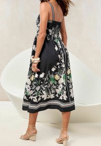 Czarna sukienka maxi w kwiaty z białymi i zielonymi wzorami, z marszczonym tyłem, cienkimi ramiączkami oraz dołem z ozdobnym wykończeniem. Noszona z sandałami na koturnie.