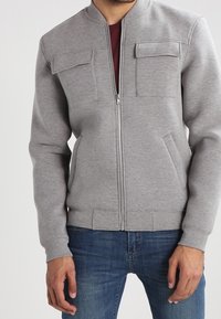 Gray zip-up sweatshirt with a round collar, two chest pockets, and ribbed cuffs. The fabric is soft and slightly textured, paired with blue jeans.