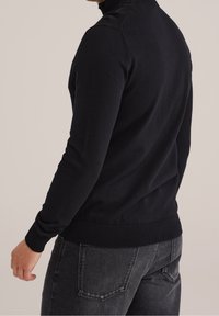 Pullover nero a maglia con colletto alto, caratterizzato da polsini e orlo a coste. Il tessuto ha una consistenza morbida e una finitura liscia. Visibili jeans neri.