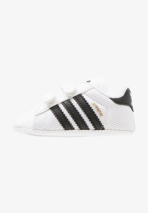 Biały sneaker Adidas Superstar z czarnymi paskami, dwoma paskami na rzepy i złotym napisem "Superstar" na boku.