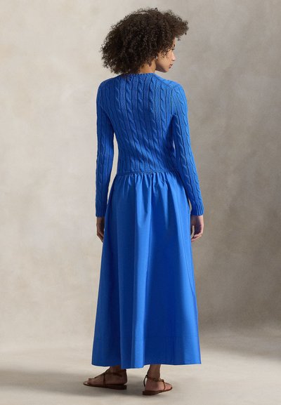 Μακρύ μπλε φόρεμα με υφή πλεκτού κορσέ και ροδαλό, αέρινο φούστα. Το ύφασμα είναι λείο, με πιέτες στη μέση. Φαίνονται σανδάλια.