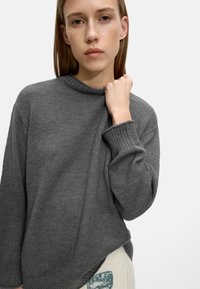 Szary sweter z dzianiny z wysokim dekoltem, prążkowanymi mankietami i luźnym krojem. Widać teksturowany materiał, w połączeniu z jasnymi spodniami.