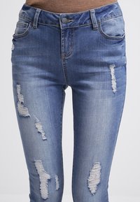 Jeans de denim azul que cuentan con un ajuste slim, detalles desgastados con acentos rotos y un diseño clásico de cinco bolsillos.