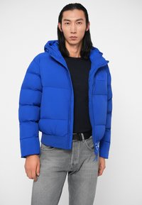 Kék puffasztott kabát kapucnival, steppelt béléssel, cipzáros záródással és bordázott mandzsettával. Fekete felső és szürke farmer alatt viselve.