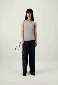 Kvinna med långt svart hår iförd en grå ärmlös topp, marinblå vida byxor, svarta skor, som håller en liten svart handväska med handledsrem.