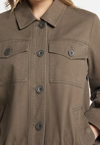 Veste brun à boutons, fabriquée en coton lisse ; elle comporte deux poches poitrine, des boutons noirs, un col pointu et des poches latérales.