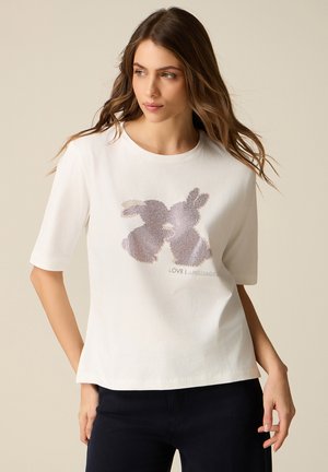 Frau mit langem Haar trägt ein weißes Hemd mit zwei lilafarbenen Hasensilhouetten und dem Schriftzug "LOVE LANGUAGE", kombiniert mit einer dunklen Hose.